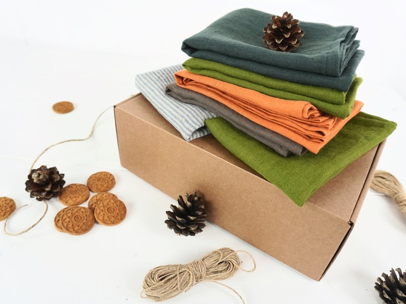 May include: A stack of different colored linen cloths in a brown cardboard box. The box is sitting on a white surface. There are pine cones and small round cookies scattered around the box.