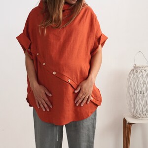 Puede incluir: Una blusa de manga corta en color naranja quemado con una tapeta de botones diagonal en la parte delantera, cuello alto y mangas con dobladillo. La prenda se combina con pantalones grises, creando un conjunto sencillo y funcional.