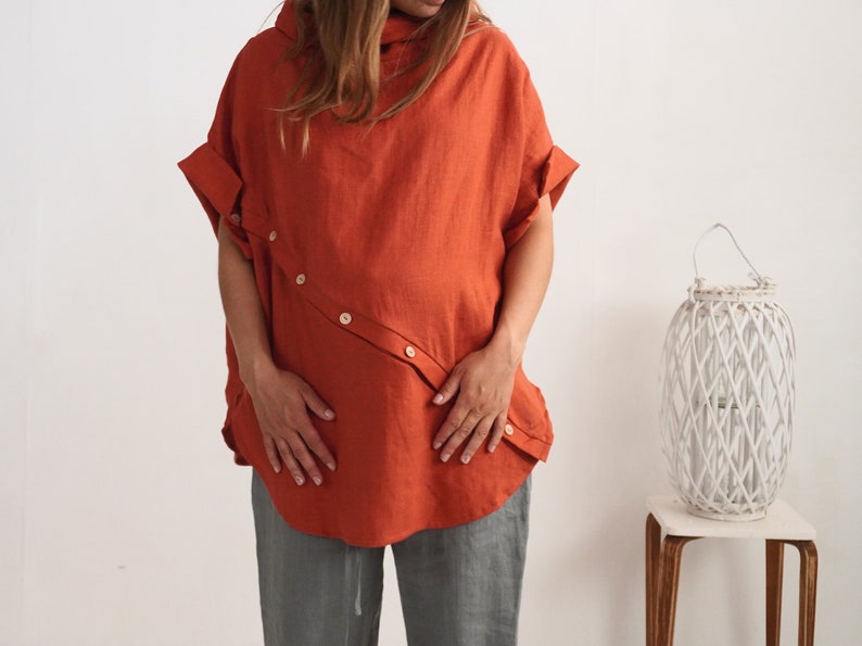 Puede incluir: Una blusa de manga corta en color naranja quemado con una tapeta de botones diagonal en la parte delantera, cuello alto y mangas con dobladillo. La prenda se combina con pantalones grises, creando un conjunto sencillo y funcional.