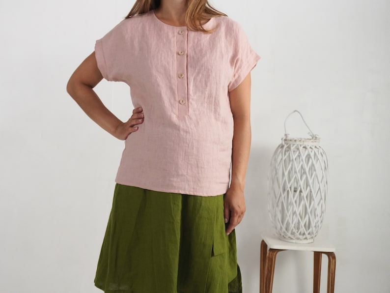 May include: A light pink short-sleeved blouse with a round neckline and a front button placket featuring four light-colored buttons, paired with a solid olive green skirt that falls to the knee, creating a simple two-piece outfit combination.