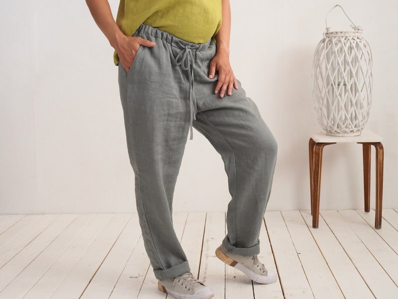 May include: Grey linen trousers with a drawstring waist and turned-up cuffs. The trousers are worn with a light green top. The model is wearing grey trainers. A white woven lantern sits on a small wooden table.