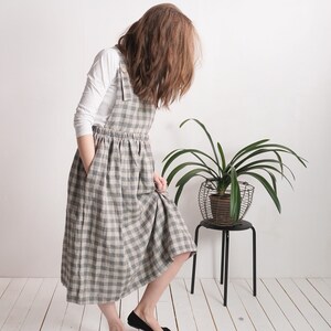 May include: A long, sleeveless pinafore dress featuring a grey and white checkered pattern. The dress has a gathered waist, side pockets, and shoulder straps with visible buttons. It is worn over a plain white long-sleeved shirt.