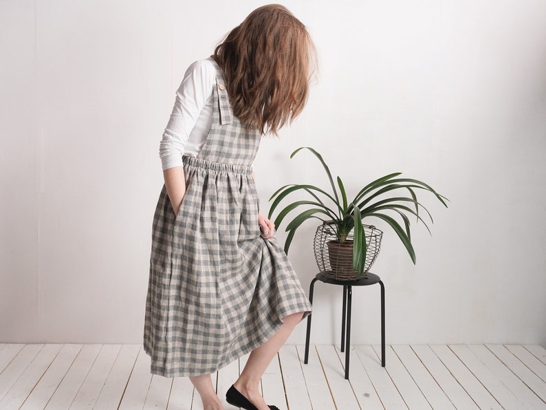 May include: A long, sleeveless pinafore dress featuring a grey and white checkered pattern. The dress has a gathered waist, side pockets, and shoulder straps with visible buttons. It is worn over a plain white long-sleeved shirt.