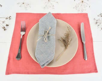 May include: A rectangular coral-colored placemat featuring a beige dinner plate, a blue and white striped linen napkin tied with twine, a silver fork, a silver knife, and a dried grass stem placed on the plate.