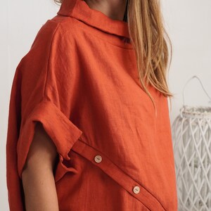Puede incluir: Una blusa de color naranja quemado con cuello alto y mangas cortas con pu&ntilde;os doblados. La prenda presenta una tapeta diagonal en la parte delantera asegurada con dos botones de madera clara, creando un aspecto estructurado.