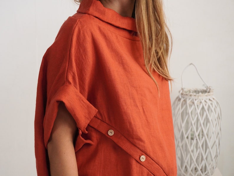 Puede incluir: Una blusa de color naranja quemado con cuello alto y mangas cortas con pu&ntilde;os doblados. La prenda presenta una tapeta diagonal en la parte delantera asegurada con dos botones de madera clara, creando un aspecto estructurado.