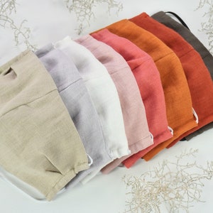 May include: A set of eight face masks made of linen fabric in various colours, including beige, grey, white, pink, orange, and brown. The masks are arranged in a stack, with the beige mask on top.