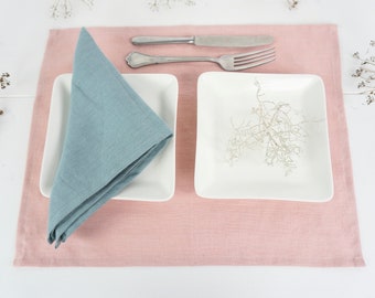 May include: A light blue linen napkin folded into a triangle on a pink linen placemat with two white square plates and silver cutlery.