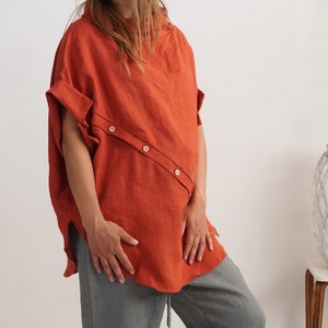 Puede incluir: Una blusa de manga corta color &oacute;xido con cuello alto y una tapeta de botones diagonal en la parte delantera con tres botones claros. La prenda tiene un corte holgado y relajado con mangas con dobladillo y bajo curvo.