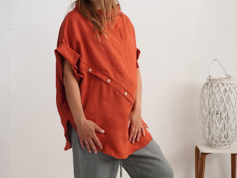 Puede incluir: Una blusa de manga corta color &oacute;xido con cuello alto y una tapeta de botones diagonal en la parte delantera con tres botones claros. La prenda tiene un corte holgado y relajado con mangas con dobladillo y bajo curvo.