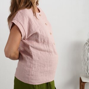May include: A light pink short-sleeved blouse featuring a round neckline and a vertical row of small, light-colored buttons down the front. The garment has cuffed sleeves and a straight hemline, paired with a solid olive green skirt.