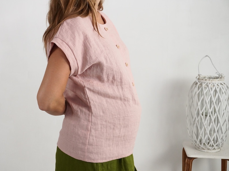 May include: A light pink short-sleeved blouse featuring a round neckline and a vertical row of small, light-colored buttons down the front. The garment has cuffed sleeves and a straight hemline, paired with a solid olive green skirt.