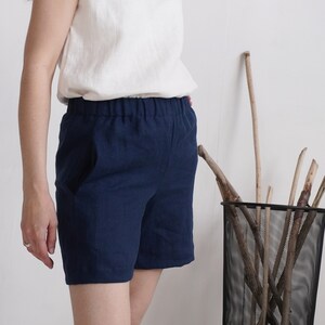 May include: Navy blue shorts featuring an elasticized waistband and side pockets. The shorts are constructed from a solid-colored fabric and reach mid-thigh length, paired with a plain white sleeveless top tucked into the waistband.