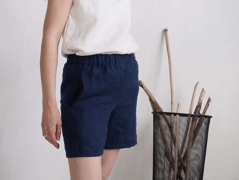 May include: Navy blue shorts featuring an elasticized waistband and side pockets. The shorts are constructed from a solid-colored fabric and reach mid-thigh length, paired with a plain white sleeveless top tucked into the waistband.