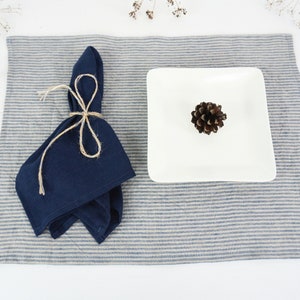May include: A blue linen napkin tied with twine on a striped placemat with a white square plate and a pine cone on top.