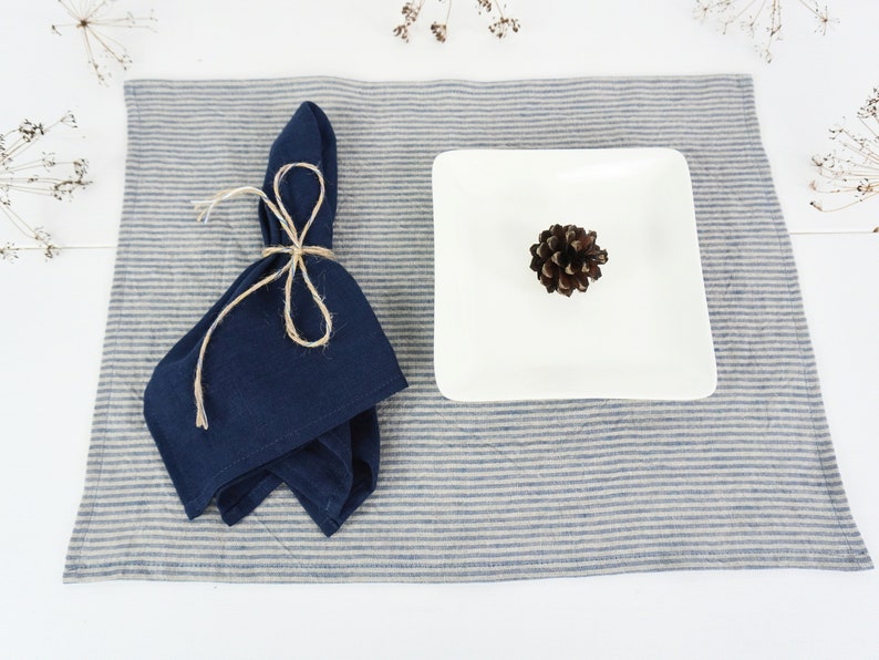 May include: A blue linen napkin tied with twine on a striped placemat with a white square plate and a pine cone on top.