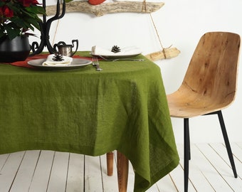 May include: A round olive green linen tablecloth draped over a wooden table. The table is set with two grey plates, white napkins topped with small pinecones, a silver fork, a black metal candelabra, and a potted poinsettia plant.
