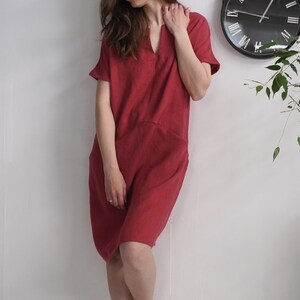 Peut inclure: Une femme portant une robe en lin rouge avec un col en V et des manches courtes. La robe est ample et a une taille l&eacute;g&egrave;rement fronc&eacute;e.