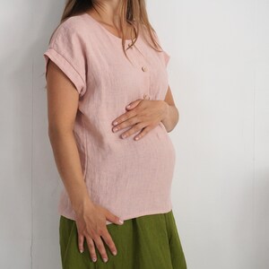 May include: A light pink short-sleeved blouse featuring a round neckline and a vertical row of three small buttons on the upper front. The garment has cuffed sleeves and a straight hemline, paired with a solid olive green skirt.