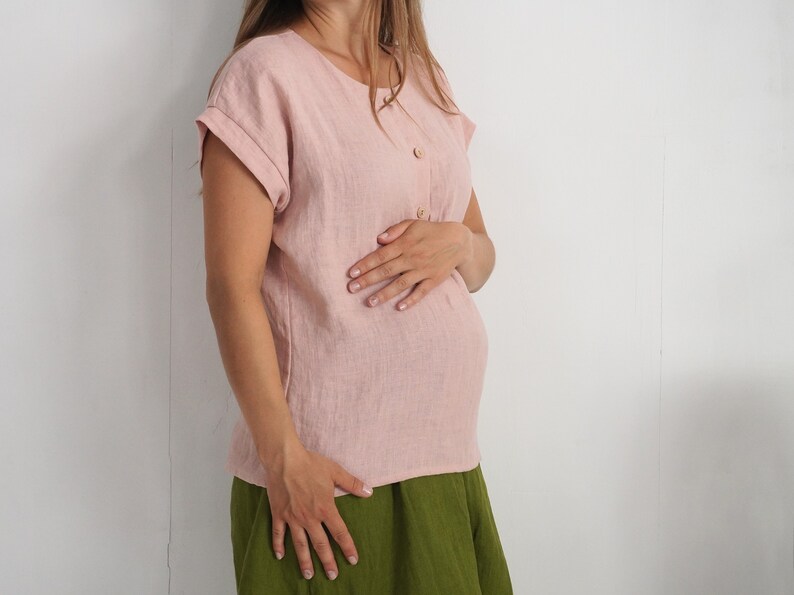 May include: A light pink short-sleeved blouse featuring a round neckline and a vertical row of three small buttons on the upper front. The garment has cuffed sleeves and a straight hemline, paired with a solid olive green skirt.