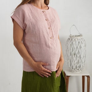 May include: A dusty pink short-sleeved blouse featuring a round neckline and a front button placket with four light brown wooden buttons. The garment has cuffed sleeves and a straight hemline, crafted from a textured fabric with visible weave.