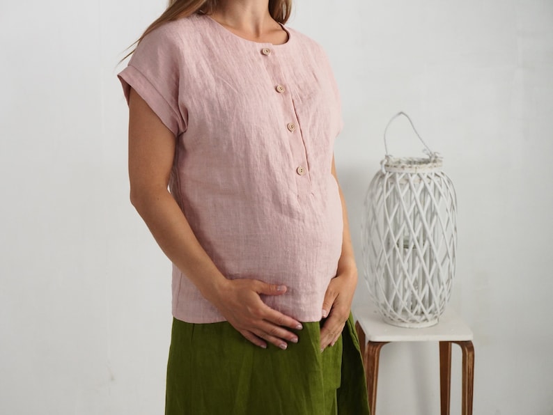 May include: A dusty pink short-sleeved blouse featuring a round neckline and a front button placket with four light brown wooden buttons. The garment has cuffed sleeves and a straight hemline, crafted from a textured fabric with visible weave.