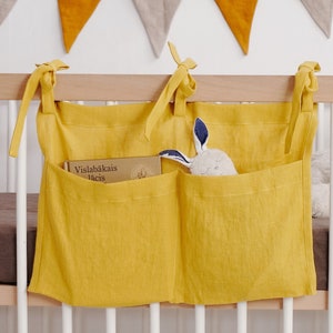 May include: A yellow linen crib organizer with three pockets. The organizer is hanging on a white crib rail. The left pocket contains a book titled "Vislabākais laiks". The middle pocket is empty. The right pocket contains a stuffed bunny.