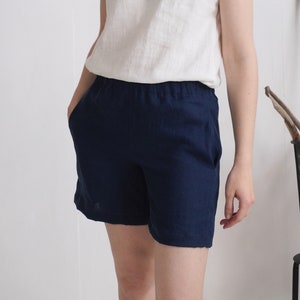 May include: Solid navy blue shorts featuring an elasticized waistband and side pockets. The shorts are paired with a plain white sleeveless top. The fabric has a visible woven texture, and the garment reaches mid-thigh with a relaxed fit.