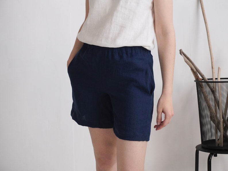 May include: Solid navy blue shorts featuring an elasticized waistband and side pockets. The shorts are paired with a plain white sleeveless top. The fabric has a visible woven texture, and the garment reaches mid-thigh with a relaxed fit.