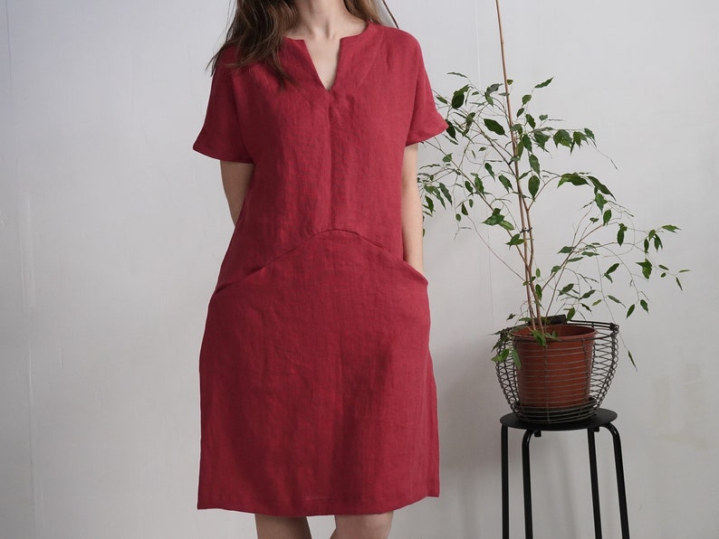 Peut inclure: Une robe en lin rouge avec un col en V et des manches courtes. La robe a deux poches sur le devant et une coupe ample.