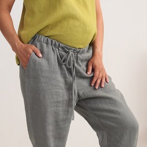 May include: Grey linen trousers with a drawstring waist and pockets. The trousers are paired with a chartreuse-coloured top. The model has their hand in the pocket of the trousers. The trousers appear to be made of a lightweight fabric.
