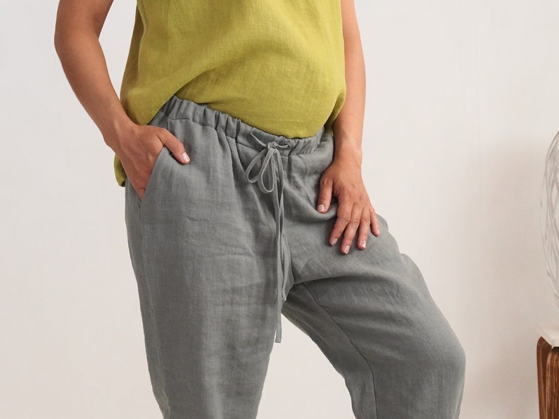 May include: Grey linen trousers with a drawstring waist and pockets. The trousers are paired with a chartreuse-coloured top. The model has their hand in the pocket of the trousers. The trousers appear to be made of a lightweight fabric.