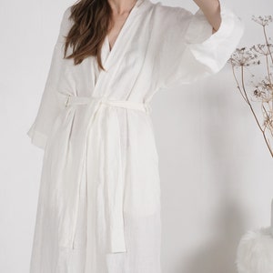 May include: A white linen robe with a long, flowing silhouette and a self-tie belt. The robe is made of a lightweight, breathable fabric and has a relaxed fit.