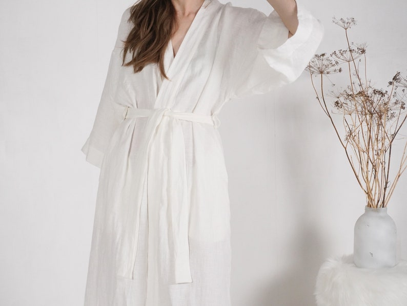 May include: A white linen robe with a long, flowing silhouette and a self-tie belt. The robe is made of a lightweight, breathable fabric and has a relaxed fit.