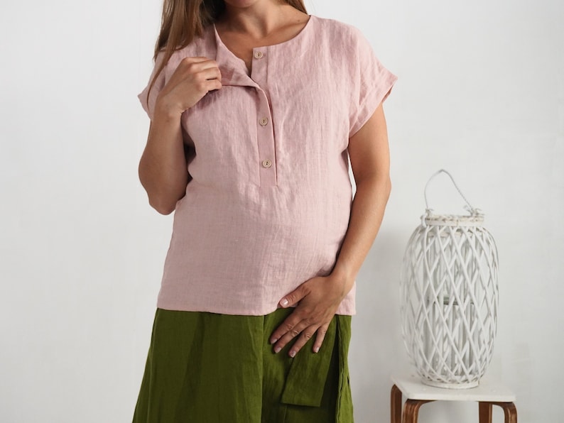 May include: A light pink short-sleeved blouse featuring a round neckline and a front placket with three visible buttons. The garment is paired with olive green trousers that have a visible fabric tie at the waist.