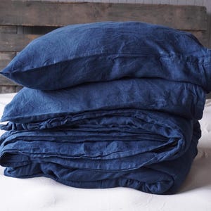 May include: A stack of blue linen bedding, including a pillowcase and a duvet cover, folded neatly on a white bed.