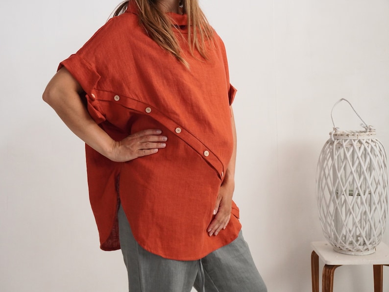 Puede incluir: Una t&uacute;nica de lino color &oacute;xido con cuello alto, mangas cortas con pu&ntilde;os doblados y una tapeta de botones diagonal que va desde el hombro hasta el dobladillo lateral. La prenda tiene un corte holgado y cuadrado con un dobladillo curvo y botones claros.