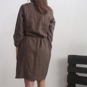 May include: A dark brown dress with long sleeves and a cinched waistline. The garment features a simple, solid-coloured design with a visible seam running down the centre back and a straight hemline that falls above the knee.