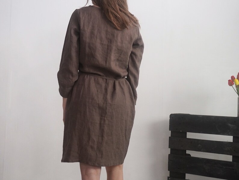 May include: A dark brown dress with long sleeves and a cinched waistline. The garment features a simple, solid-coloured design with a visible seam running down the centre back and a straight hemline that falls above the knee.