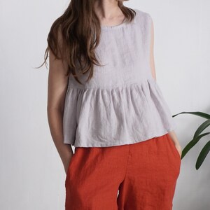 Puede incluir: Una blusa sin mangas de color lavanda claro con cuello redondo y cintura con volante fruncido, combinada con pantalones holgados de color naranja quemado. La blusa presenta un dise&ntilde;o sencillo con un acabado de tela texturizada y silueta fluida.