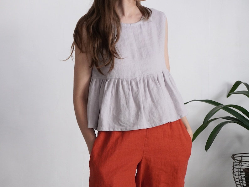 Puede incluir: Una blusa sin mangas de color lavanda claro con cuello redondo y cintura con volante fruncido, combinada con pantalones holgados de color naranja quemado. La blusa presenta un dise&ntilde;o sencillo con un acabado de tela texturizada y silueta fluida.