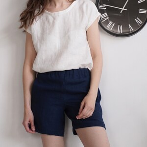 May include: A white short-sleeved top with a round neckline paired with navy blue shorts featuring an elastic waistband. The outfit is presented against a plain white wall with a black circular wall clock visible in the upper right corner.