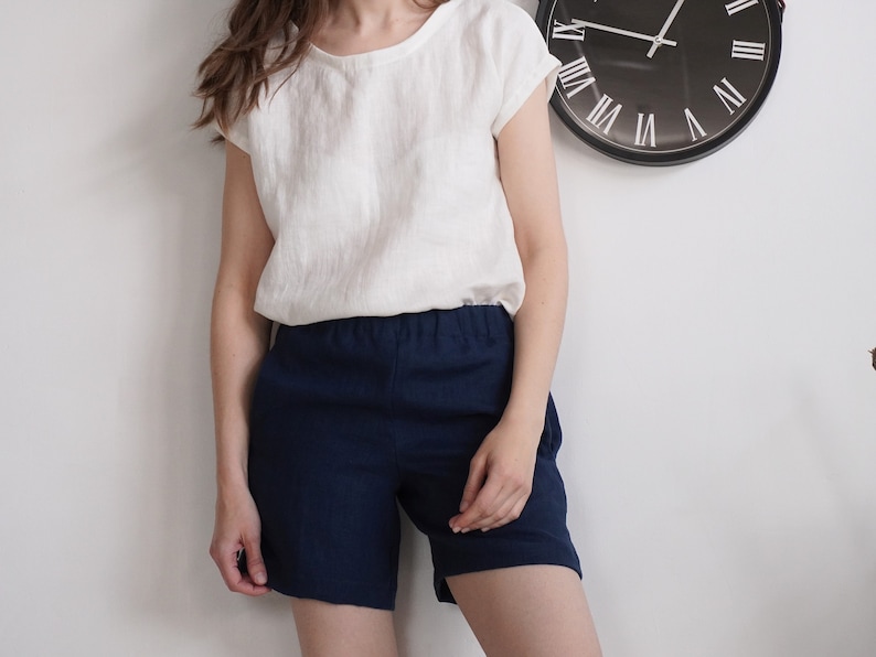 May include: A white short-sleeved top with a round neckline paired with navy blue shorts featuring an elastic waistband. The outfit is presented against a plain white wall with a black circular wall clock visible in the upper right corner.