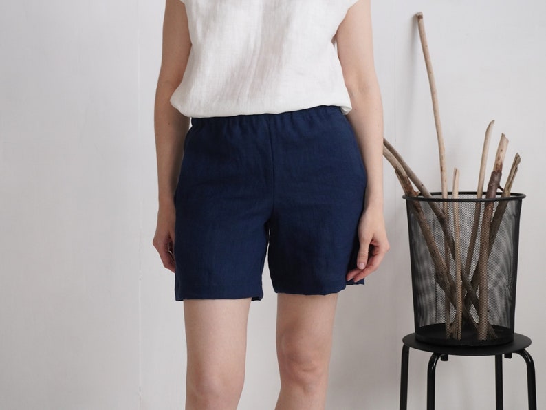 May include: A pair of dark navy blue shorts with an elasticized waistband, paired with a simple white sleeveless top. The shorts feature a clean, straight-leg cut that reaches mid-thigh, crafted from a textured fabric with visible weave details.