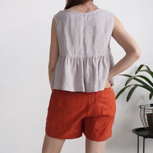Puede incluir: Una blusa sin mangas de color lavanda claro con cintura p&eacute;plum fruncida y cuello redondo, combinada con pantalones cortos de color naranja quemado. La blusa presenta una costura visible en la parte media, mientras que los pantalones cortos tienen dos bolsillos traseros y un dobladillo recto.