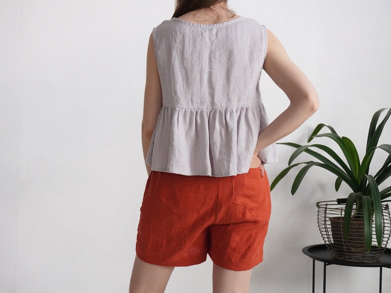 Puede incluir: Una blusa sin mangas de color lavanda claro con cintura p&eacute;plum fruncida y cuello redondo, combinada con pantalones cortos de color naranja quemado. La blusa presenta una costura visible en la parte media, mientras que los pantalones cortos tienen dos bolsillos traseros y un dobladillo recto.