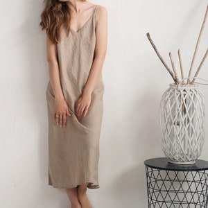 Op de afbeelding: Een beige mouwloze midi-jurk met een V-hals en dunne spaghettibandjes. De jurk heeft een los, recht silhouet dat tot halverwege de kuit valt en is gemaakt van een gestructureerde stof in een neutrale, aardse beige kleur.