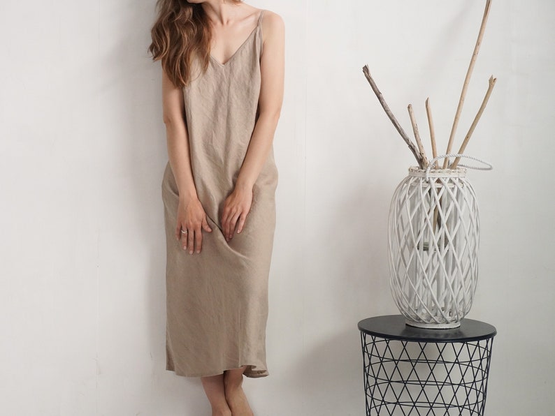 Op de afbeelding: Een beige mouwloze midi-jurk met een V-hals en dunne spaghettibandjes. De jurk heeft een los, recht silhouet dat tot halverwege de kuit valt en is gemaakt van een gestructureerde stof in een neutrale, aardse beige kleur.