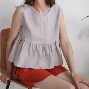 Puede incluir: Una blusa sin mangas de color lavanda claro con cuello redondo y cintura con volante fruncido. La prenda presenta un acabado de tela texturizada y se combina con pantalones cortos de color naranja quemado liso que llegan a medio muslo.