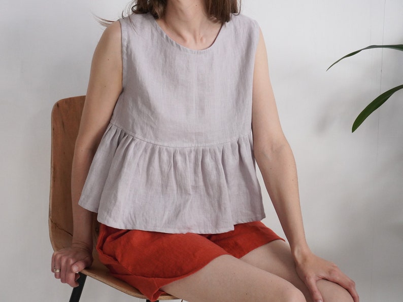 Puede incluir: Una blusa sin mangas de color lavanda claro con cuello redondo y cintura con volante fruncido. La prenda presenta un acabado de tela texturizada y se combina con pantalones cortos de color naranja quemado liso que llegan a medio muslo.